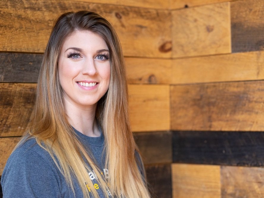 Headshot photo of Amy Duff, A long haired female, white with brown hair wearing a gray t-shirt standing in front of a wood slat background wall