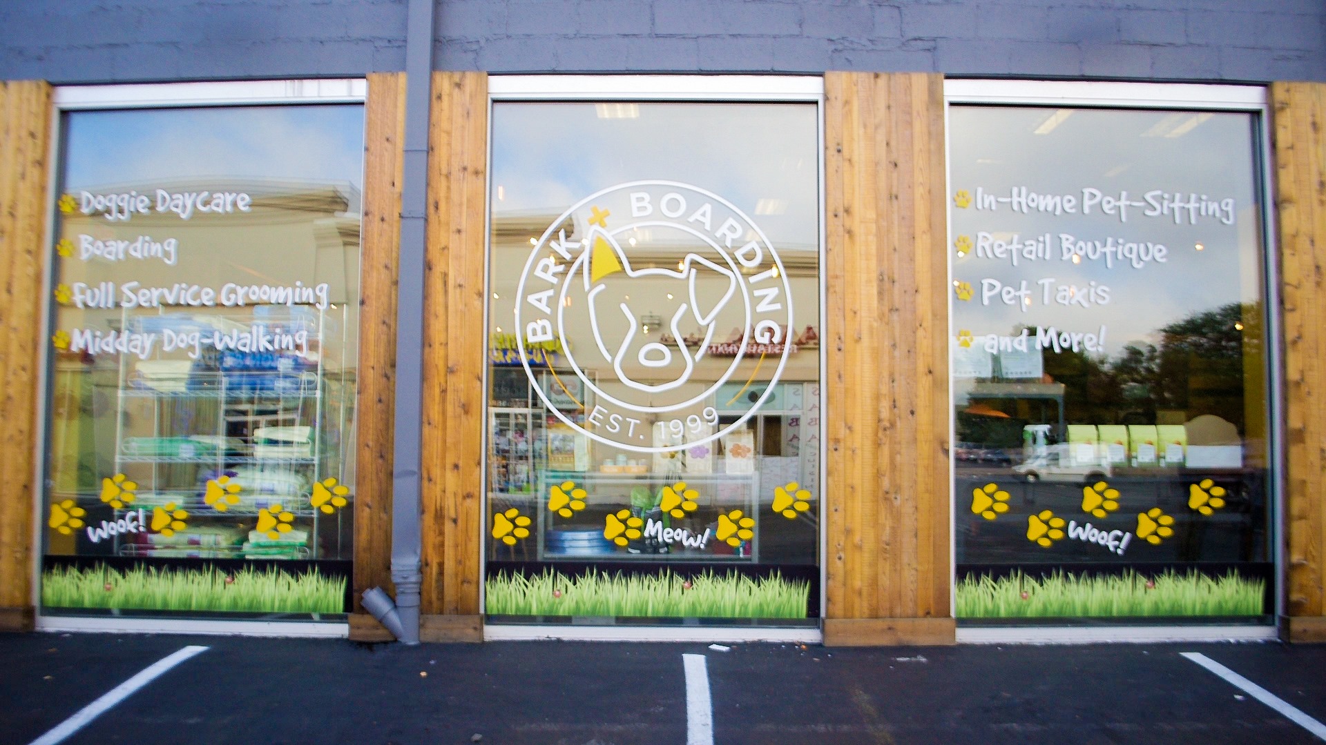 Exterior image of Bark + Boarding. 3 windows seperated by wood panels with gray industrial colored details. the center window pane has the bark + boarding logo. the left panel lists services including doggie daycare, boarding, full service grooming, and midday dog-walking. the right panel lists services including: in-home pet-sitting, retail boutique, pet taxis, and more! There are yellow paw prints along the bottom of all three windows and green pet friendly grass at the bottom of each window.